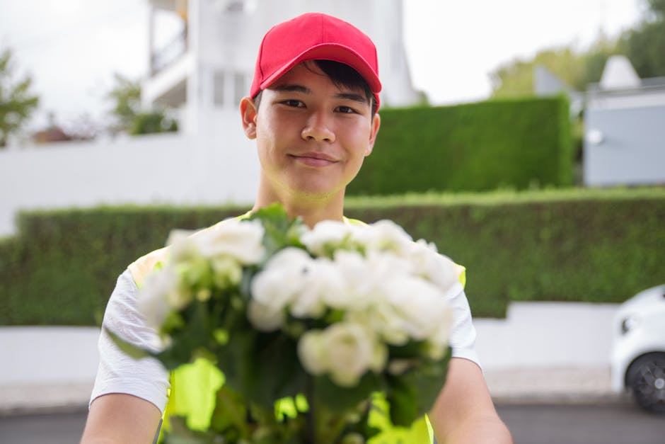 A young male flower delivery courier wearing a red cap and a high-visibility vest extends his hands towards the camera, holding a vibrant bouquet of fresh white roses and green foliage. The bouquet is