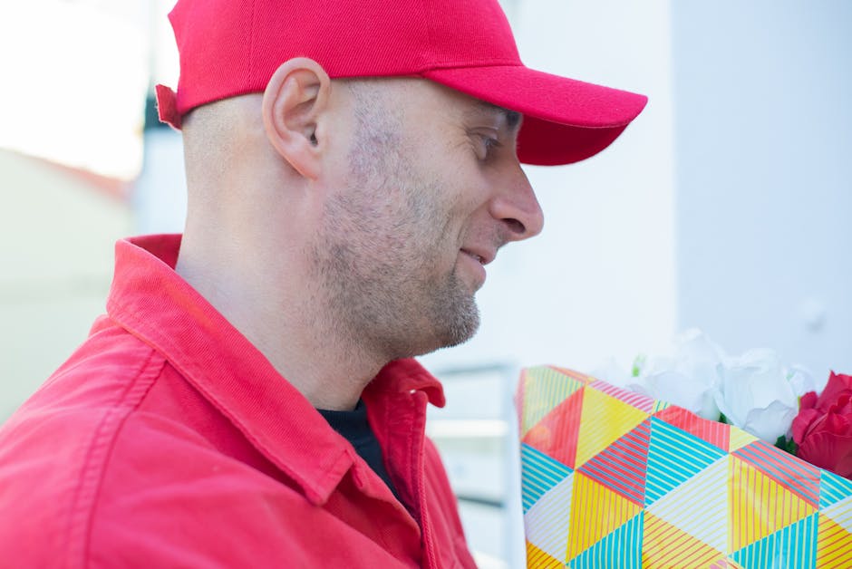 A delivery man wearing a red cap and red jacket holding a floral gift box with a colorful geometric pattern. The gift box contains fresh flowers, including white and pink blooms, wrapped in white tiss