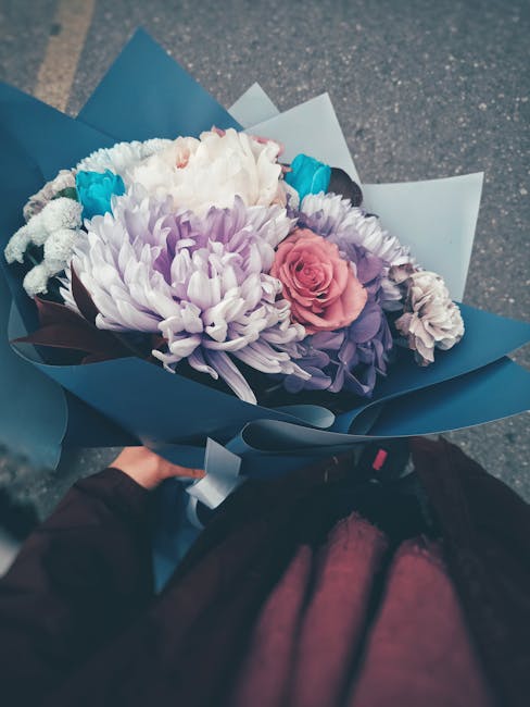A bouquet of fresh flowers being held by a person, featuring a large white chrysanthemum, soft pink roses, purple lisianthus, and bright blue accents, all arranged with lush green foliage. The arrange