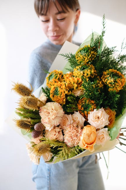A person holding a large floral bouquet featuring a mix of fresh flowers, including soft peach roses, vibrant yellow and orange marigolds, pale pink carnations, and textured green foliage, wrapped in
