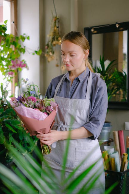 A young woman with fair skin and light brown hair tied back, dressed in a light blue shirt and a white apron, holding a fresh bouquet of flowers in her hands. The bouquet features pink and purple flow
