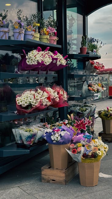 An outdoor display of various floral arrangements and bouquet arrangements arranged on dark metal shelves at a flower shop, featuring a variety of fresh flowers including white daisies, pink lilies, a