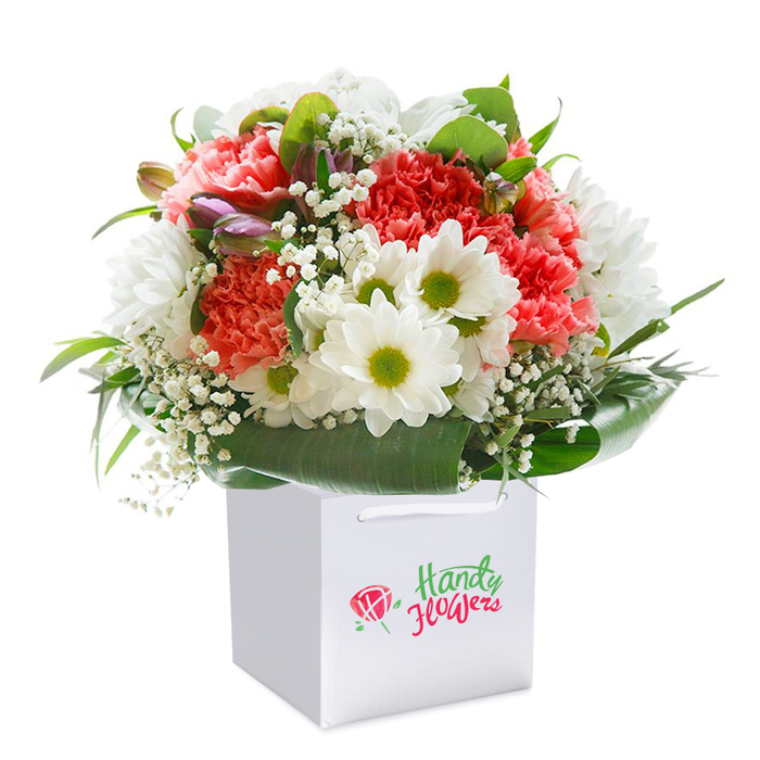 Settled gracefully in a sharply squared white gift bag, a stunning floral composition stands as a testament to skilled floristry in Hillingdon. The bouquet's central focus lies in the interplay of lush coral and peach carnations, their frilly petals splayed in vibrant profusion, evoking the gentle colour palette of summer gardens along Long Lane. Dotted generously amongst them are snowy daisy-like blooms, each white ray circling a chartreuse center bright with joy-calling to mind leisurely afternoons by the River Pinn. Delicate baby's breath is threaded throughout, providing a feathery, cloud-like contrast to the solid color blocks, while on the left, several tightly furled buds-tinged purple and promising future bloom-lend a quiet intrigue that deepens the arrangement. Crisp, broad leaves in deep, resinous green curve around the base, offering sturdy structure and a rich frame as glossy as the foliage in Minet Country Park. The bouquet is rounded, full, and fresh, with petals catching the soft, diffused light that accentuates the intricate layering and attention to detail. White ribbon handles crown the bag, ready for carrying to an event in Yiewsley or for doorstep delivery to a loved one in Hillingdon Heath. The elegant script and rose emblem printed on the bag's front quietly underscore the quality and local credibility of the presentation, making this refined gift perfect for a birthday, anniversary, or simple reassurance sent from heart to home, anywhere across Hillingdon.
