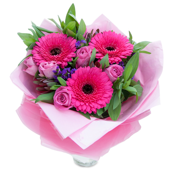 I picture this bouquet placed on a café table in Hillingdon on a soft afternoon: three vivacious fuchsia Gerbera daisies open wide, each petal band catching the light while deep, almost black centres provide a striking counterpoint. Around them, dusty-rose roses in a delicate lavender-pink are still gently unfurling, their tender curves offering a softer rhythm to the arrangement. Tiny clusters of purple statice thread through the composition like confetti, their papery texture creating little pockets of contrast and a sense of movement. The greenery is generous - broad salal-like leaves and elongated eucalyptus pick up the cool tones of the roses and add a refreshing, dewy aroma that complements the faint sweetness of the gerberas. The bouquet is beautifully hand-wrapped: an inner cushion of matte pale pink tissue supports the stems while an outer sheet of translucent fuchsia-pink cellophane, slightly crinkled, adds a shimmering finish and reinforces the colour story. Carefully folded, the wrap reveals the top rim of a clear glass vase below, suggesting the arrangement is ready to brighten a windowsill or dining table. Lighting here is soft and even, allowing the varied textures - from the velvet rose petals to the crisp foliage and glossy cellophane - to read clearly. As a local florist I imagine delivering this to a neighbour near Hillingdon Court Park or leaving it as a surprise on a friend's doorstep: it's celebratory yet intimate, a perfect choice for birthdays, Mother's Day, or as a small, thoughtful gesture that carries warmth.