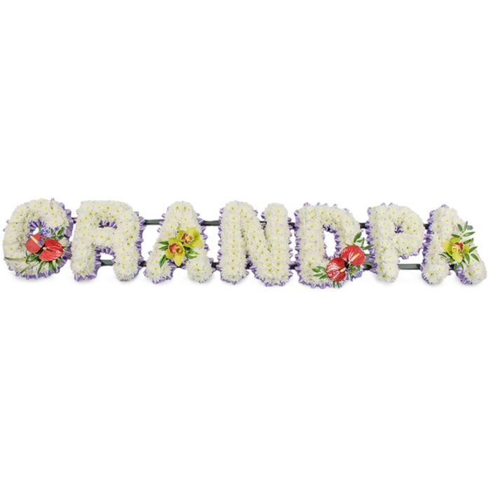 A carefully crafted floral arrangement stretches horizontally to spell the word "GRANDPA" in solid capital letters, designed as a memorial tribute for a much-loved grandfather in Hillingdon. Each character is fully covered with pristine white chrysanthemums, the blooms arranged so closely that no gaps of foam or framework are visible. This deliberate density creates a plush, almost quilted appearance, inviting the eye to follow the word from left to right. A band of soft pastel lavender ribbon traces the base of each letter, subtly visible beneath the flowers and giving a gentle lilac glow that outlines the shapes, adding just enough colour to stop the white from feeling stark. The letters rest on a concealed structure of dark green bars, which provide stability while blending into the foliage elements. Four accent clusters bring warmth and personality to the tribute. The "G" and the central "D" each carry a grouping of deep red anthuriums, their smooth, waxy, heart-shaped spathes arching outward, with bright yellow spadices standing proudly at the centre. These are surrounded by lush green leaves and small hints of purple filler flowers, suggesting both devotion and remembrance. The first and last "A" display sprays of yellow-lime orchids, likely cymbidium, with broad, velvety petals and gently speckled throats, framed by glossy greenery that softens the edges. The clean white background and even lighting allow every petal and ribbon edge to be seen clearly, as though the tribute has just been finished in a local Hillingdon flower shop before being sent to a funeral at a nearby church or crematorium. The result is a bespoke GRANDPA tribute that feels dignified, loving, and deeply personal.